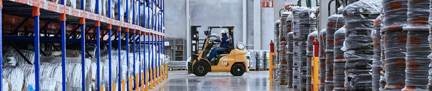 Forklift operator navigating through a busy manufacturing warehouse aisle stocked with pallet racking and wrapped raw material coils.