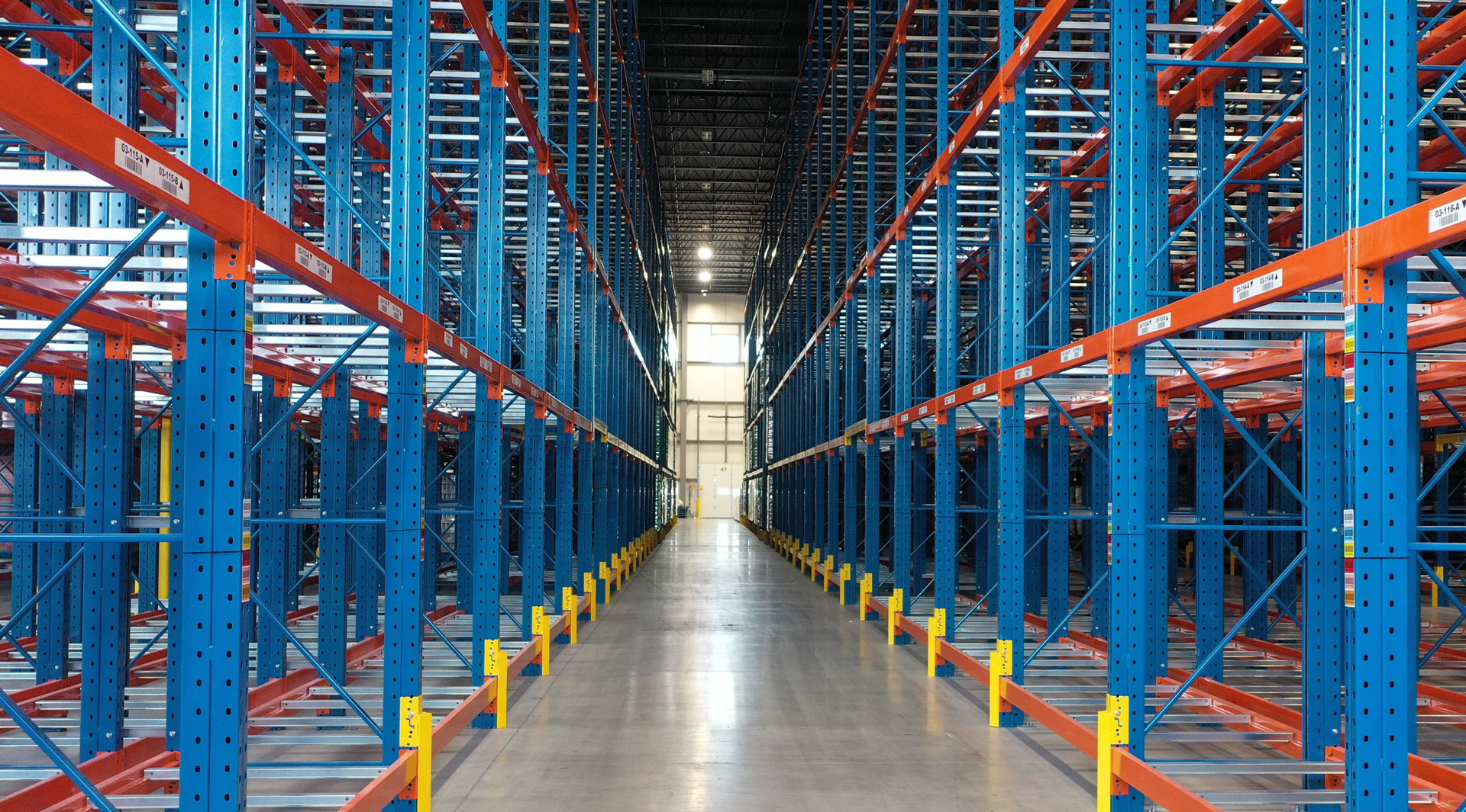 Symmetrical, eye-level view looking down a central aisle surrounded by blue and orange double deep pallet racking with yellow base guards.