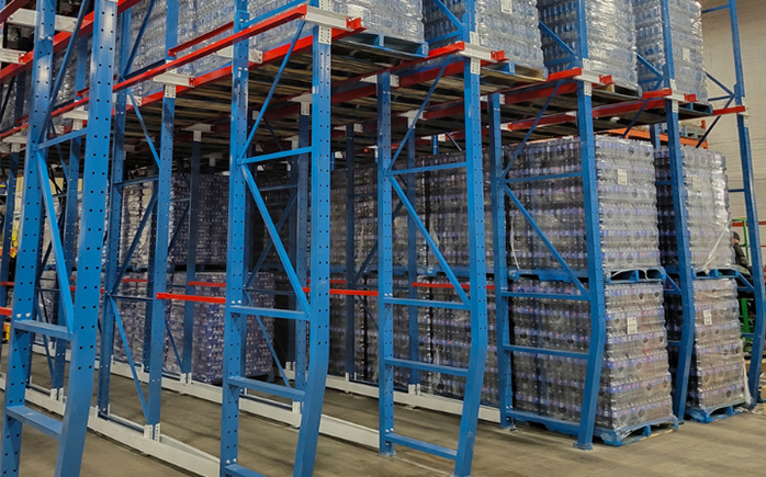 High-density blue drive-in pallet racking system with red support rails fully loaded with shrink-wrapped beverage pallets.