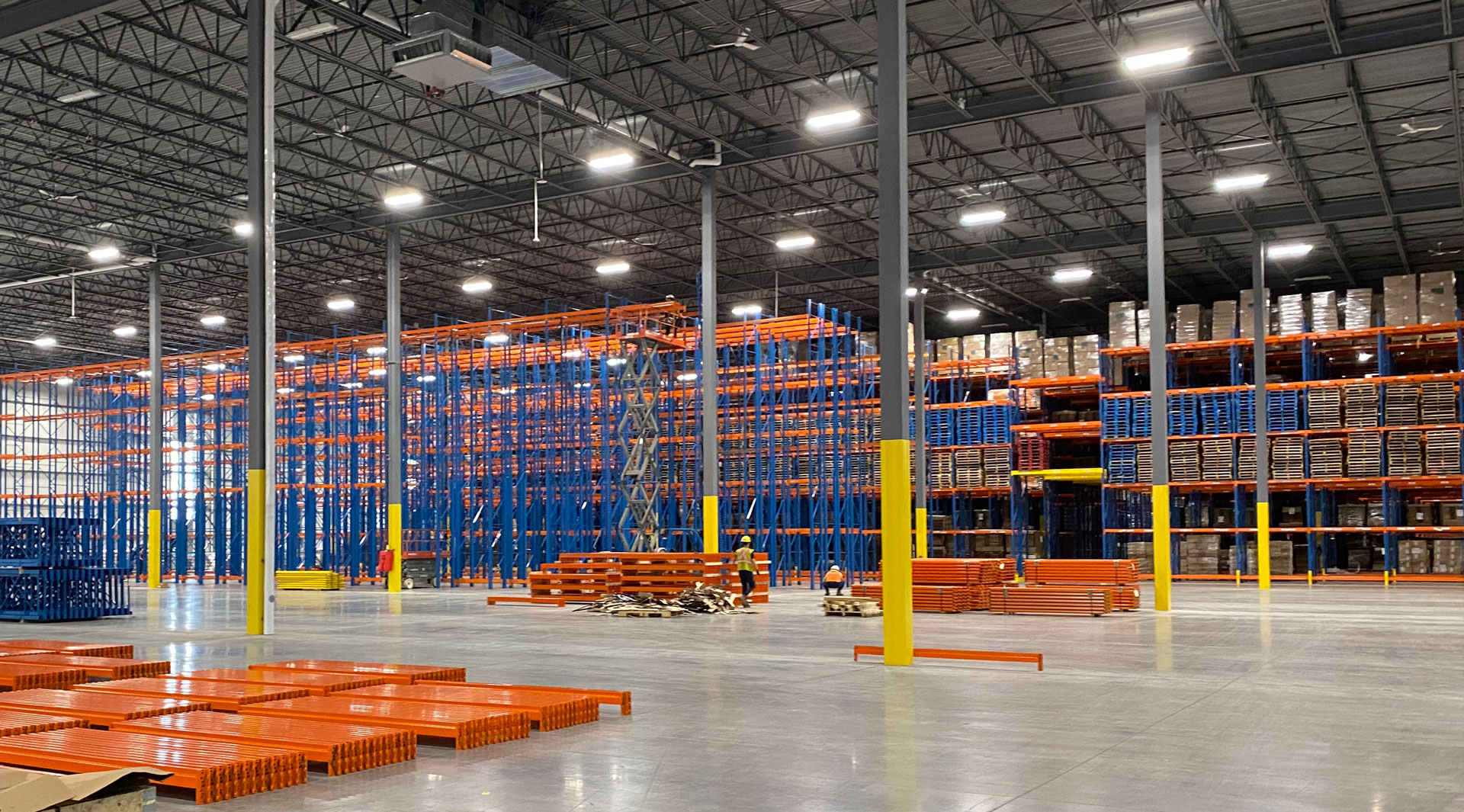 Wide view of a large-scale warehouse installation featuring blue uprights, orange beam components stacked on the floor, and a worker on a scissor lift.