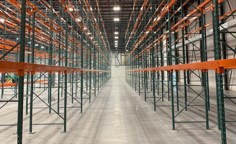 Symmetrical, ground-level view looking down a wide aisle between tall rows of empty green and orange selective pallet racking.