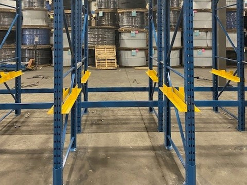 Front-facing aisle view of a blue cable reel racking system with yellow axle brackets and large wire spools stored in the background.