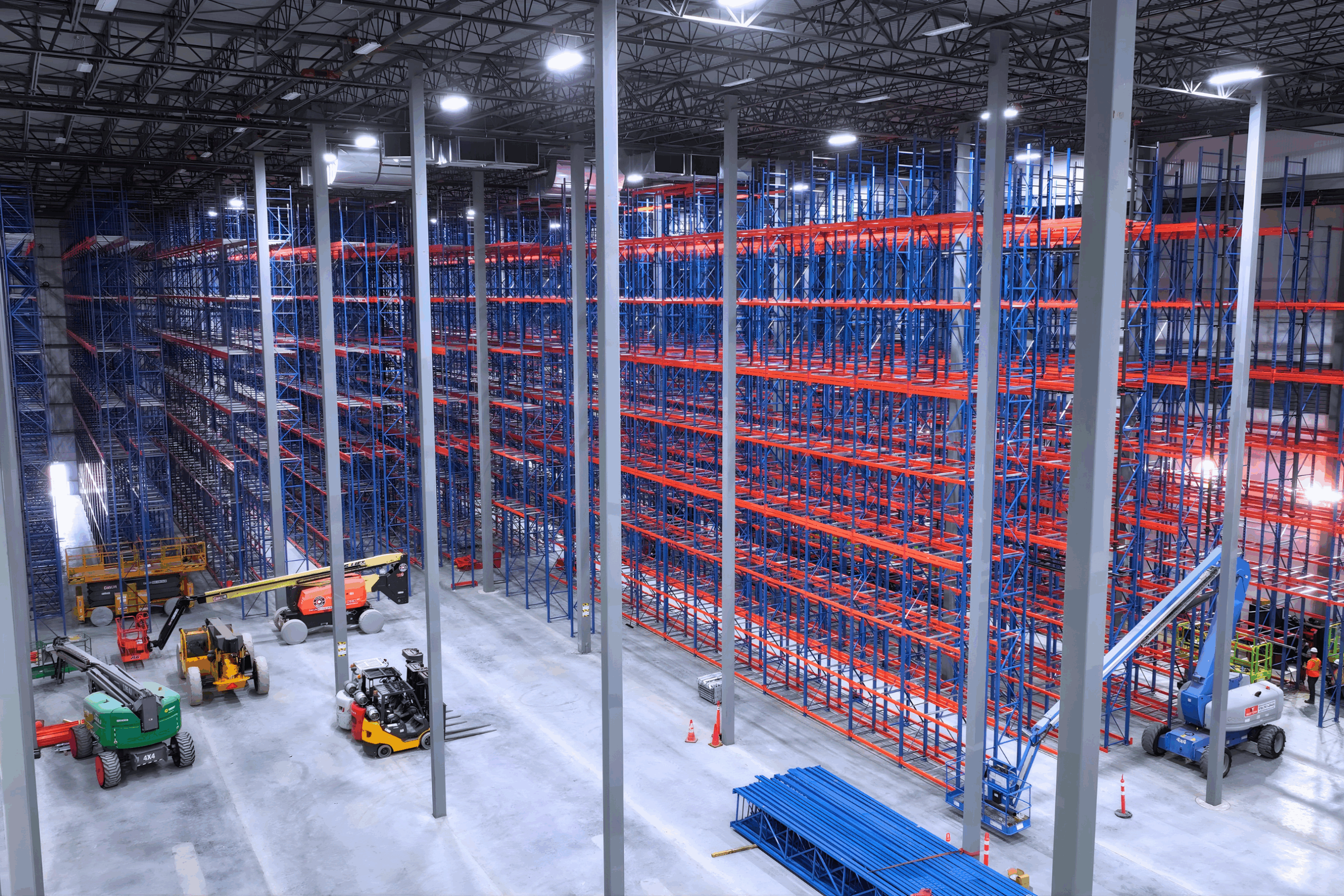 High-angle wide shot of a massive warehouse under construction featuring a blue and orange Very Narrow Aisle (VNA) racking system, with multiple aerial lifts and forklifts on the concrete floor