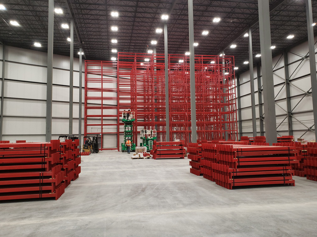 Installation stage of a tall red VNA selective racking system with workers operating scissor lifts and organized stacks of rack components in the foreground.