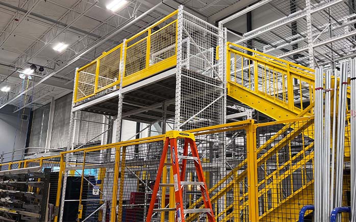 Angle view of an industrial pick tower module showing an upper walkway platform, yellow stair system, and wire mesh security partitions.