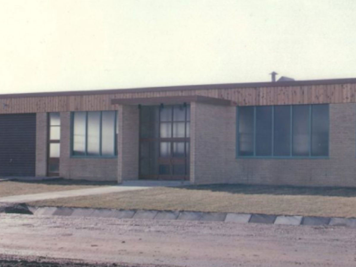 Vintage exterior photograph of a single-story commercial building featuring light brick, wood panel trim, and glass front doors.
