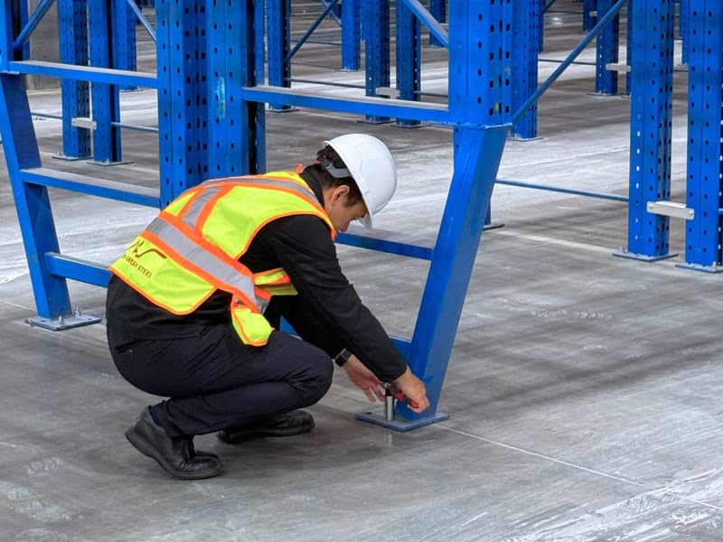 Certified engineer crouching down to inspect and adjust the floor anchor bolts on a blue pallet rack upright column baseplate.