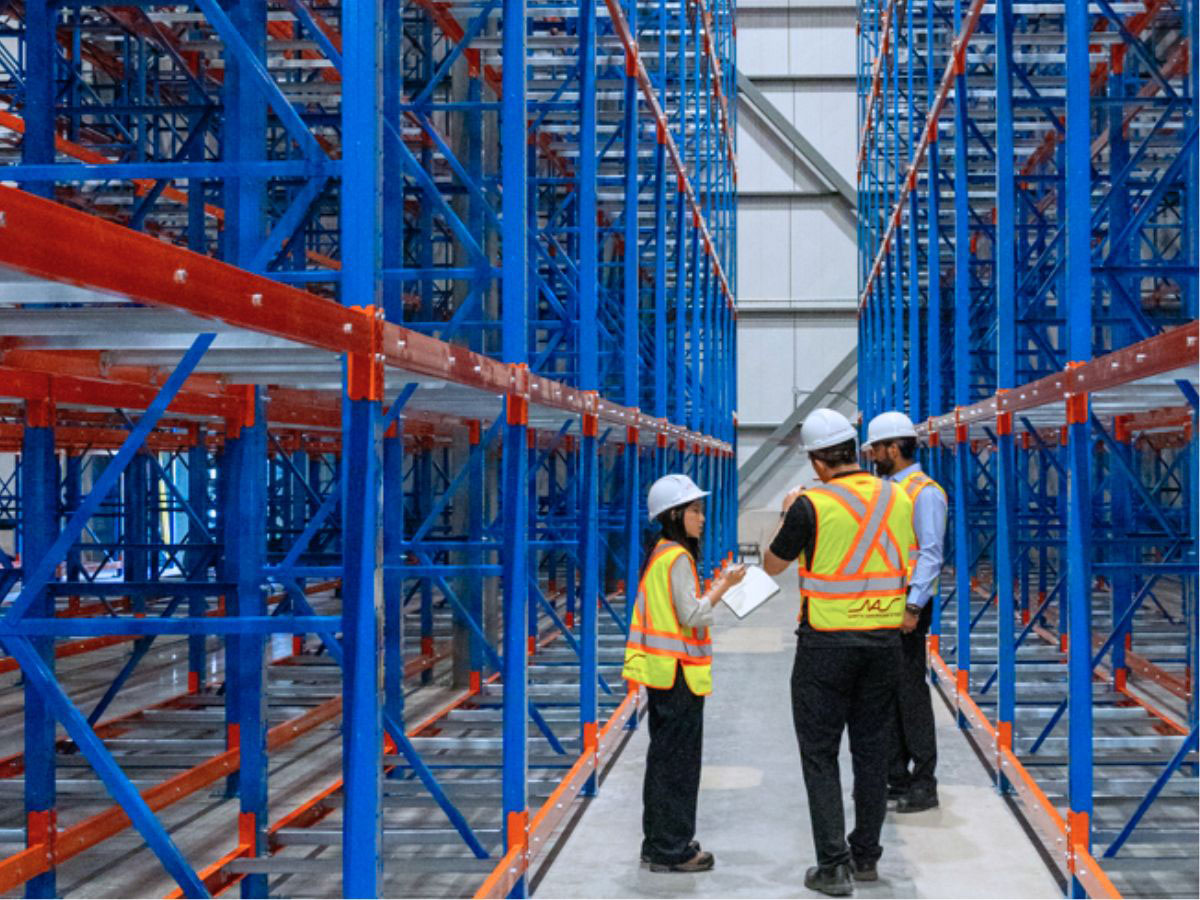 Industrial inspection view of three warehouse workers in high-visibility vests and hard hats conducting an audit inside an aisle framed by blue uprights and orange beam structural selective pallet racking.