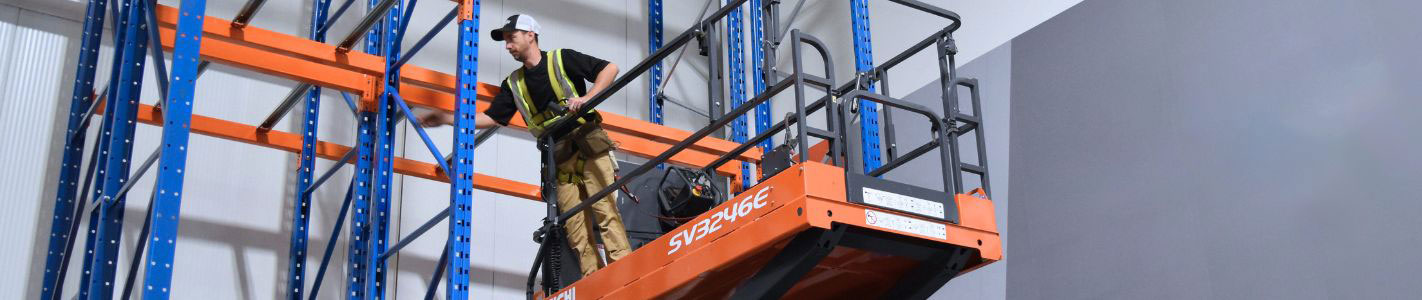 Warehouse installer on an orange scissor lift assembling high-density blue pallet racking uprights and orange cross beams.