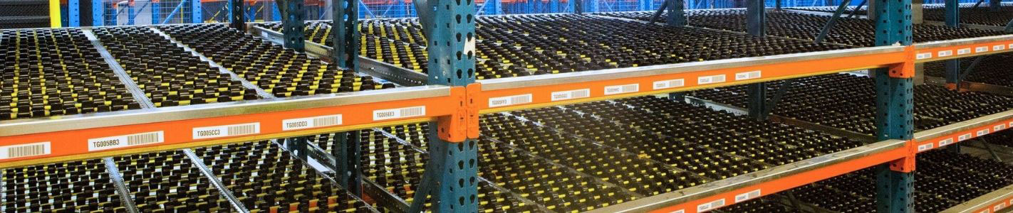 An inclined carton flow rack system loaded with dense rows of inventory bottles, framed by blue uprights and orange shelf beams with barcode labeling.