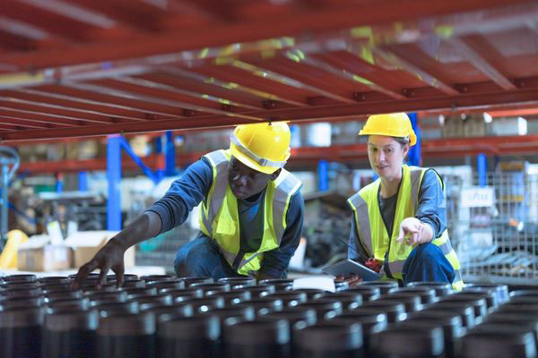 Two warehouse employees wearing hard hats and safety vests inspecting small parts stored in a gravity flow picking module.