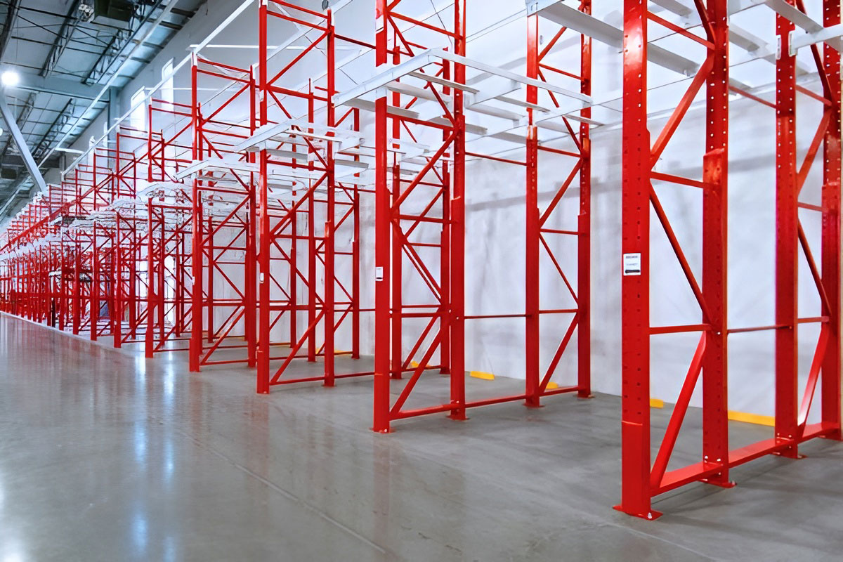 Side-angle view of empty red structural steel drive in racking frames installed along a concrete warehouse floor.