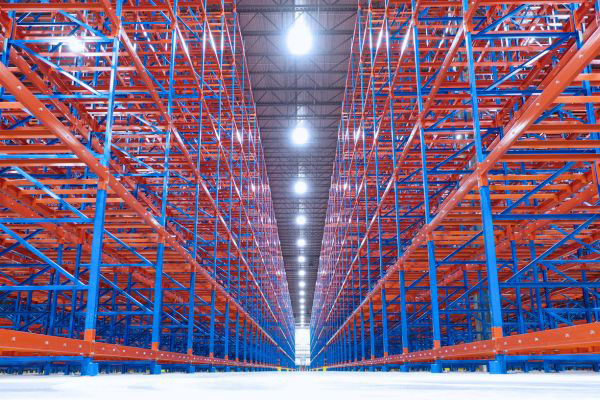 Symmetrical low-angle view looking down a central aisle of tall, empty blue and orange pallet racking in a large warehouse.