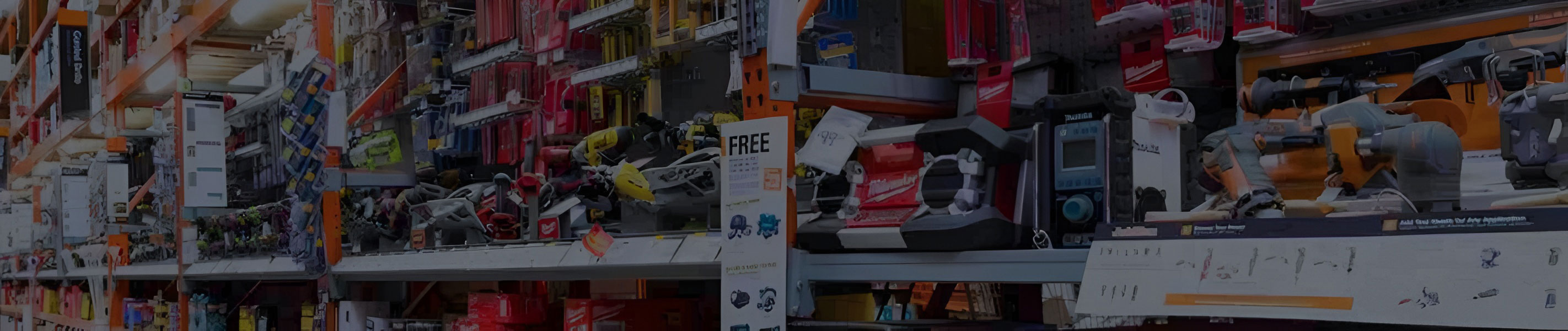 A retail hardware store aisle with orange industrial beam shelving densely stocked with boxed power tools, accessories, and packaged store merchandise.