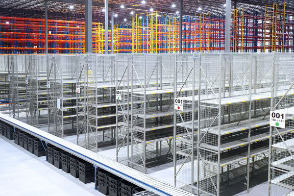 Elevated view of organized grey steel industrial shelving units with mesh side panels and aisle markers alongside a roller conveyor line.