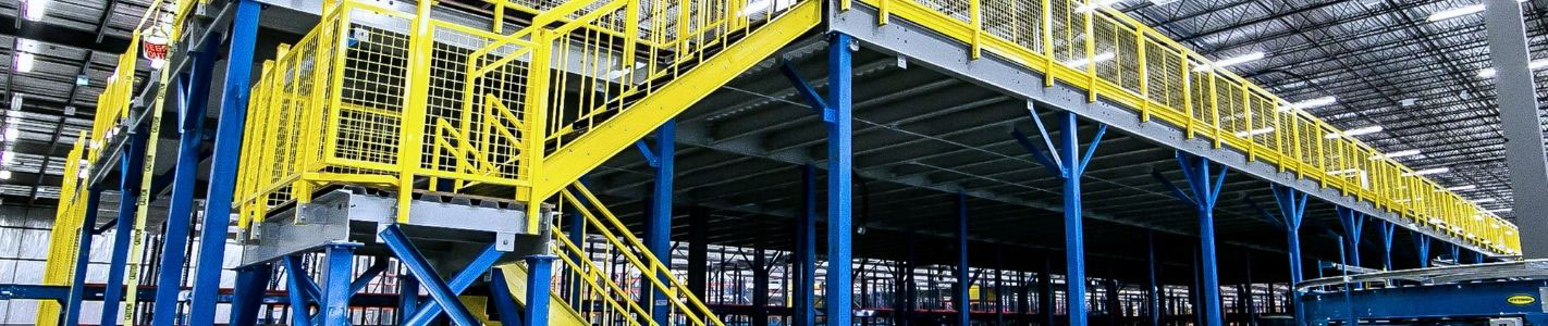 Structural steel mezzanine platform in a high-ceiling warehouse, supported by blue vertical columns and equipped with bright yellow safety handrails and a perimeter staircase.